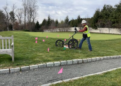 A worker wearing hardhat and reflective vest pushing a scanning equipment with three large wheels. The ground also has orange flag markings.